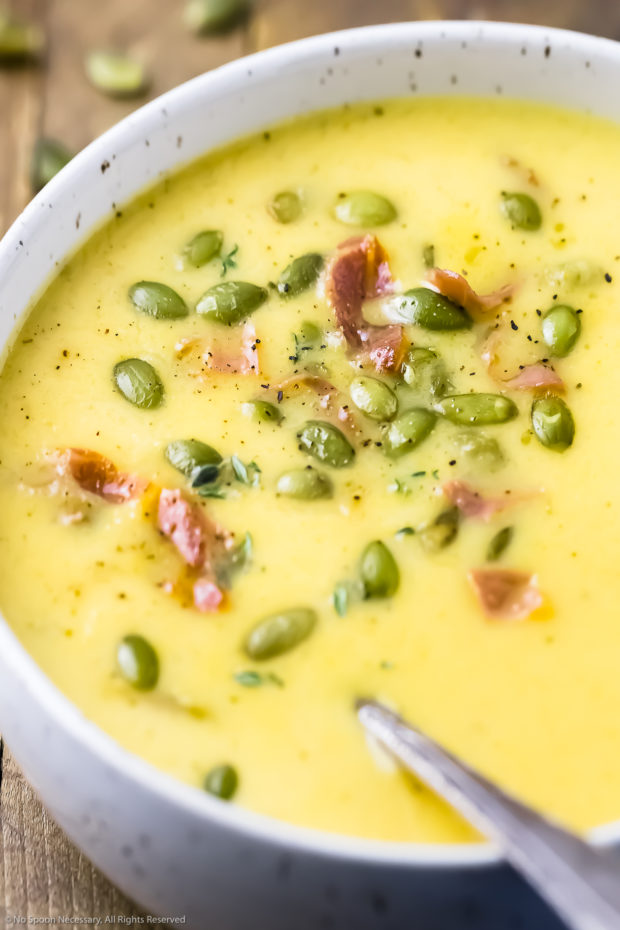 Angled, close-up photo of a bowl of Apple Soup garnished with pumpkin seeds and crispy bacon with a spoon inserted in to the soup.