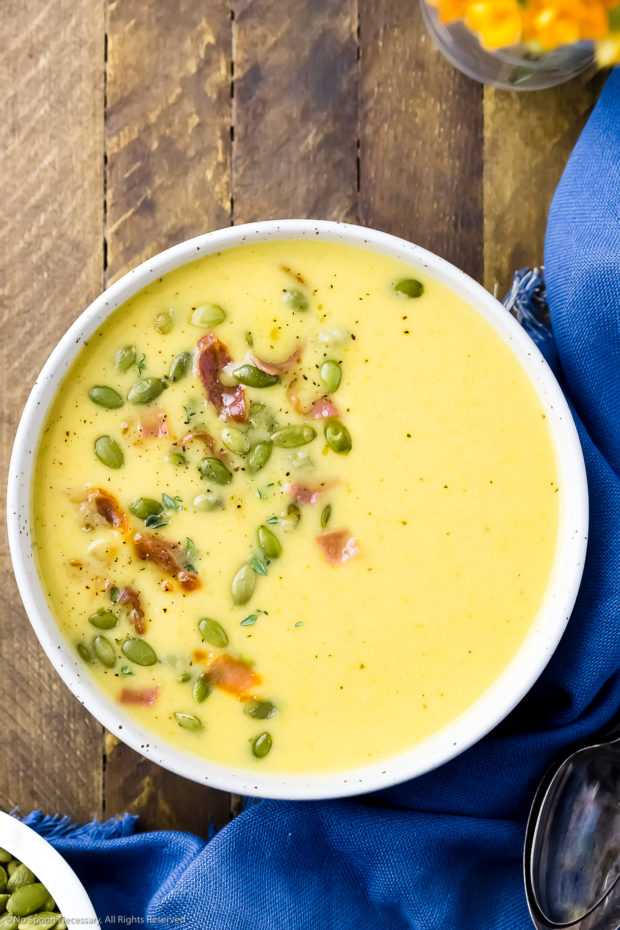 Overhead shot of a bowl of Cheddar Apple Soup garnished with pumpkin seeds and crispy bacon with a blue linen, spoons and orange flowers in a vase surrounding the bowl.