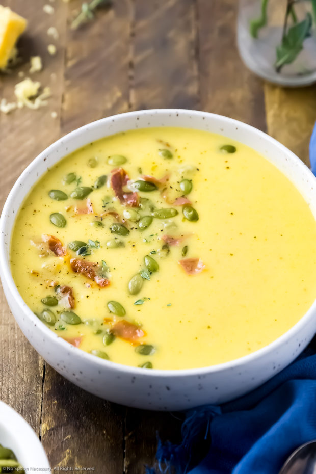 Angled photo of a bowl Apple Cheddar Cheese Soup with potatoes garnished with pumpkin seeds and crispy bacon with a blue linen next to the bowl and a block of cheddar blurred in the background.