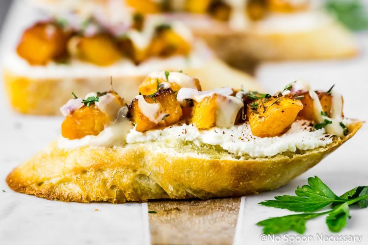 Straight on shot of a Caramelized Butternut Squash Crostini topped with whipped Feta, za'atar and tahini on a marble and wood bread board with more crostini blurred in the background.