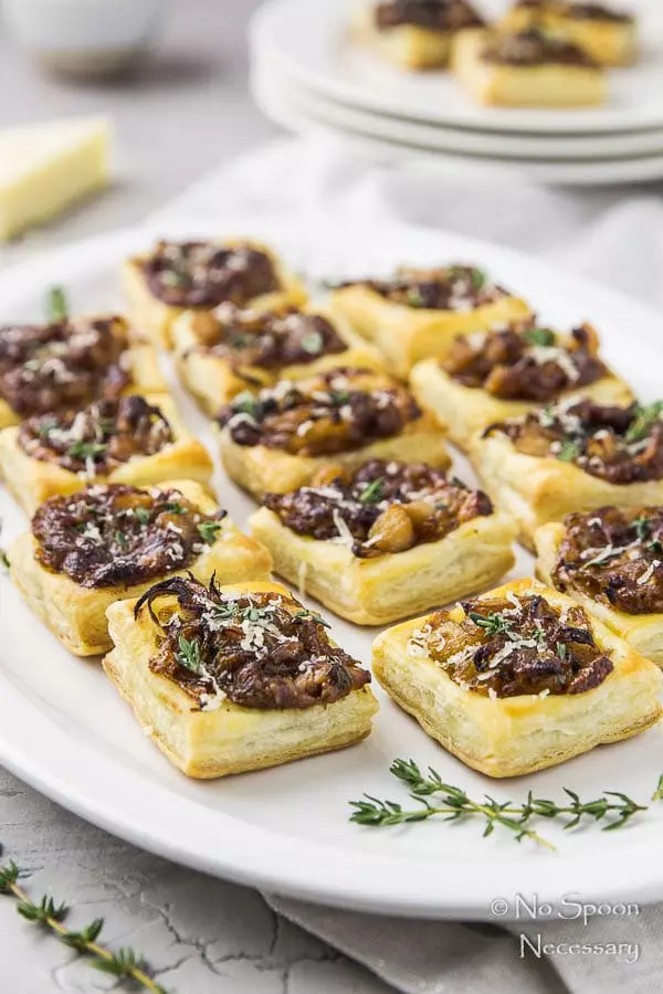 Angled shot of a white platter filled with Caramelized Onion Pear Tarts and fresh thyme with a block of gorgonzola and stack of plates with more tarts blurred in the background.