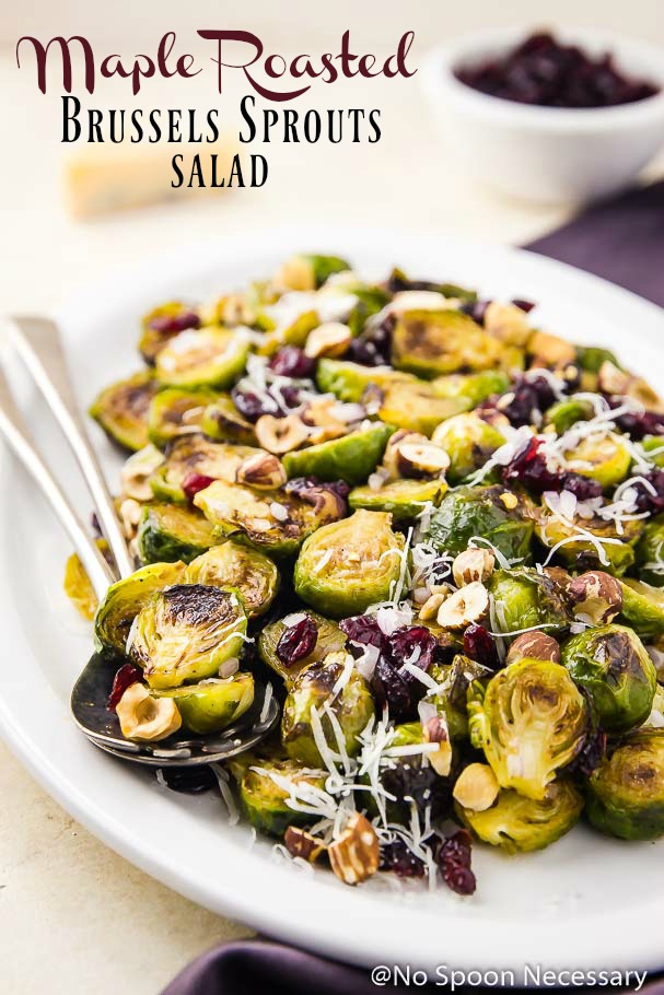Angled shot of a white platter of Maple Roasted Brussels Sprouts Salad with with shredded pecorino, hazelnuts, and dried cranberries; with serving spoons tucked under the brussels sprouts and a ramekin of cranberries blurred in the background
