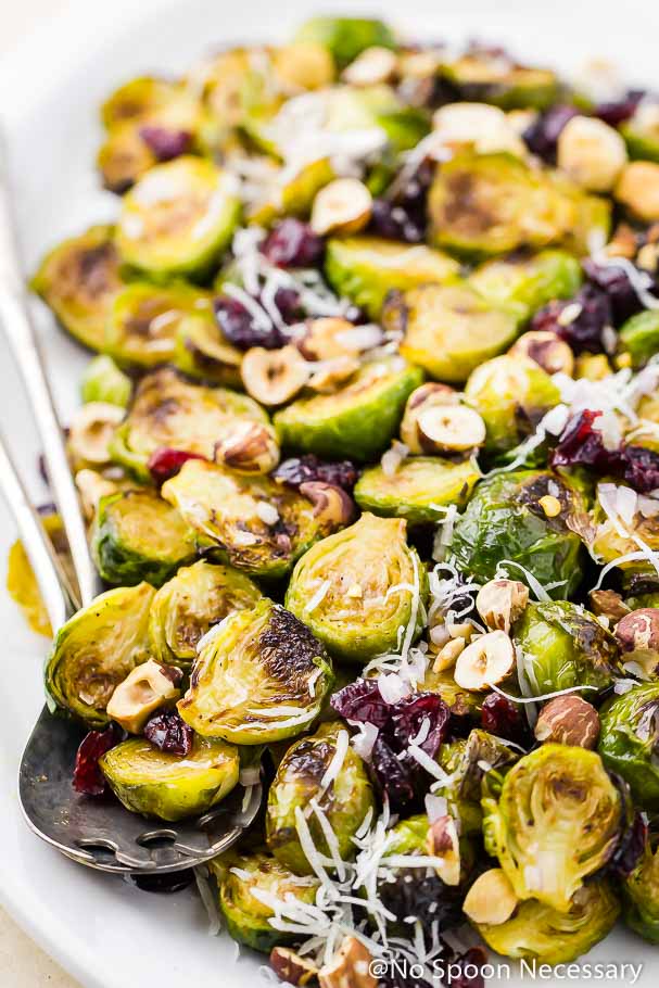 Angled shot of a white platter of Maple Roasted Brussels Sprouts Salad with shredded pecorino, toasted hazelnuts and dried cranberries with serving spoons tucked under the brussels sprouts