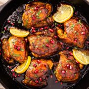 Overhead photo of pomegranate chicken in a cast-iron skillet with pomegranate molasses sauce and lemon wedges.