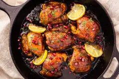 Overhead photo of pomegranate chicken in a cast-iron skillet with pomegranate molasses sauce and lemon wedges.