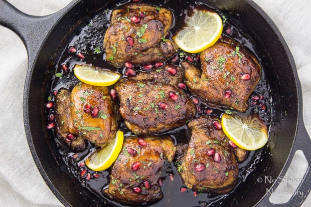 Overhead shot of Skillet Honey Pomegranate Chicken Thighs in a cast iron skillet garnished with lemon wedges and pomegranate arils with a light neutral linen under the skillet.