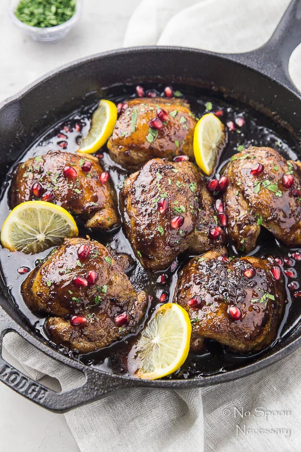 Angled shot of Skillet Honey Pomegranate Chicken Thighs in a cast iron skillet with lemon wedges and pomegranate arils with a light neutral linen under the skillet and a ramekin of fresh parsley blurred in the background. 