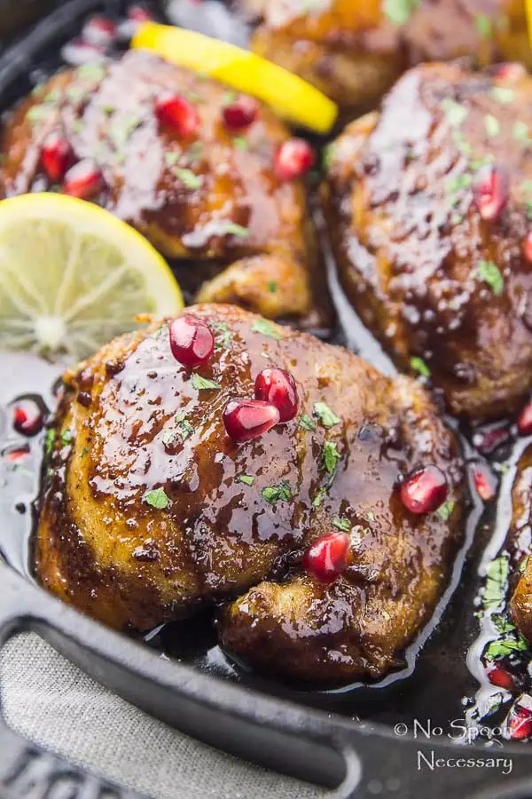 Angled, up close shot of Skillet Honey Pomegranate Chicken Thighs in a cast iron skillet garnished with lemon wedges, fresh parsley and pomegranate arils.