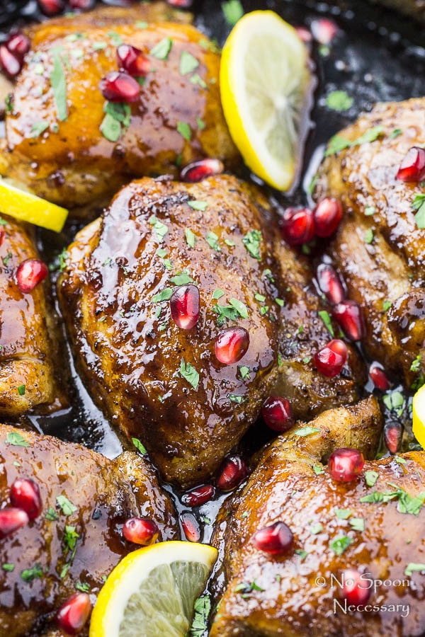 Overhead, up close shot of Skillet Honey Pomegranate Chicken Thighs in a cast iron skillet with lemon wedges and pomegranate arils with the focus on one individual chicken thigh.