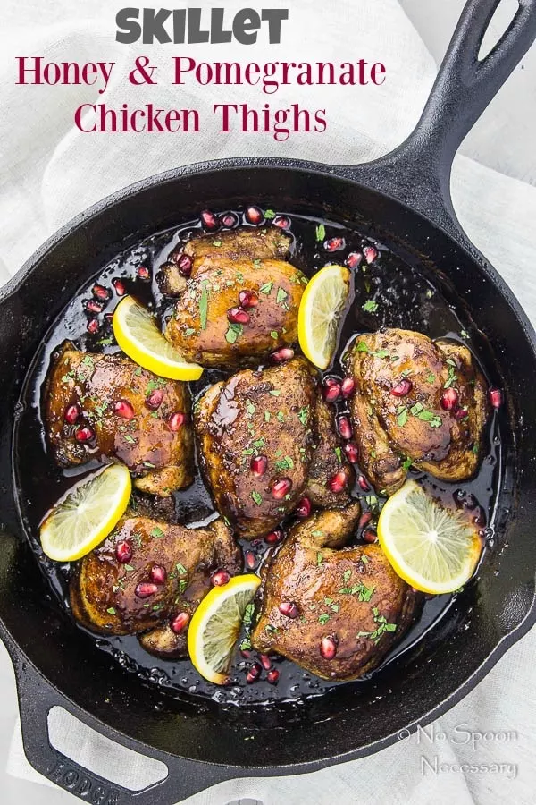 Overhead shot of Skillet Honey Pomegranate Chicken Thighs in a cast iron skillet with lemon wedges and pomegranate arils with a light neutral linen under the skillet.