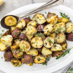 Overhead shot of Asiago, Garlic & Herb Roasted Red Potatoes on a white platter with serving spoons tucked under the potatoes; a small block of parmesan and fresh herbs in the corner