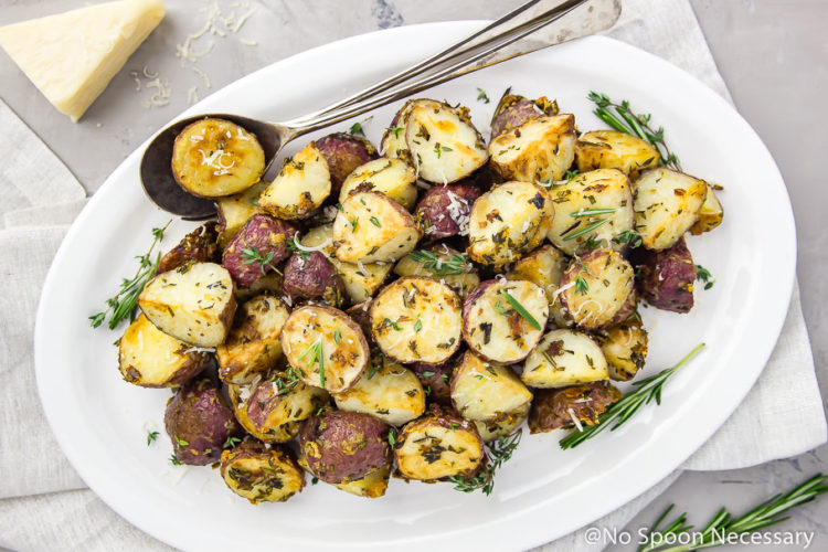 Overhead shot of Asiago, Garlic & Herb Roasted Red Potatoes on a white platter with serving spoons tucked under the potatoes; a small block of parmesan and fresh herbs in the corner