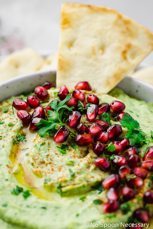 45 degree angle, up close shot of Holiday Hummus (arugula and roasted garlic white bean hummus) garnished with fresh parsley and pomegranate arils in a white bowl with a pita triangle dipped in