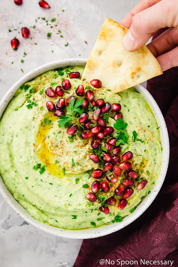 Overhead shot of a hand with a pita chips dipping into Holiday Hummus (arugula and roasted garlic white bean hummus) garnished with fresh parsley and pomegranate arils in a white bowl with a deep purple linen on the side of the bowl