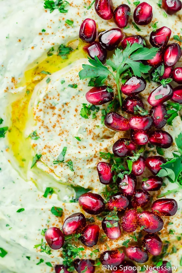 Overhead, extreme up close shot of Holiday Hummus (arugula and roasted garlic white bean hummus) garnished with a drizzle of olive oil, fresh parsley and pomegranate arils