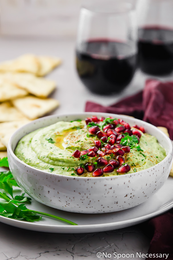 Straight on shot of Holiday Hummus (arugula and roasted garlic white bean hummus) garnished with fresh parsley and pomegranate arils in a white bowl on a white plate with pita triangles, fresh parsley stems, a deep purple linen and two red wine glasses blurred in the background
