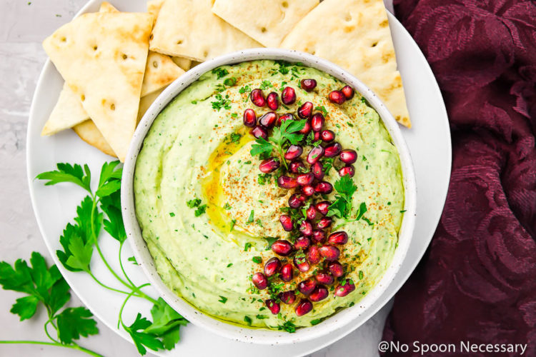 Overhead, landscape shot of Holiday hummus (arugula and roasted garlic) garnished with fresh parsley and pomegranate arils in a white bowl on a white plate with pita triangles and fresh parsley stems with a deep purple linen on the side.