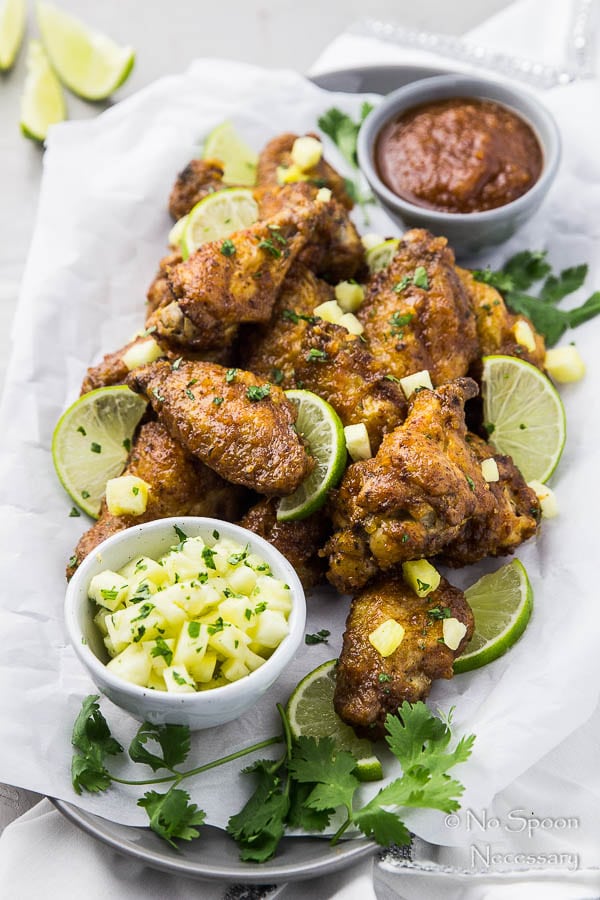 45 degree angle shot of Baked Crispy Cajun Chicken Wings garnished with pineapple salsa on a parchment paper lined platter with two small bowls of honey chipotle glaze and salsa with lime wedges and fresh parsley scattered around the platter.