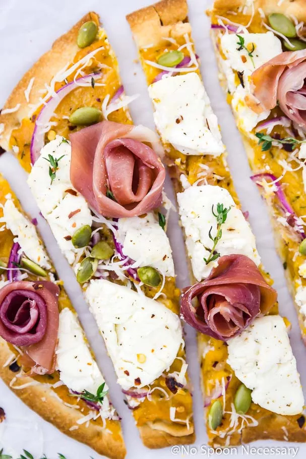 Overhead, up-close shot of a few long vertical slices of Butternut Squash Flatbread Pizza with Goat Cheese, Prosciutto & Pumpkin Seeds on a crinkled piece of parchment paper