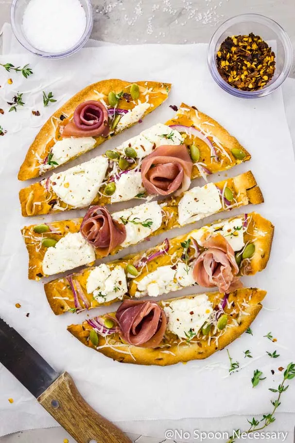 Overhead shot of Butternut Squash Flatbread Pizza with Goat Cheese, Prosciutto & Pumpkin Seeds cut into long horizontal slices on a crinkled piece of parchment paper with a wood handled steak knife, fresh thyme, ramekin of salt and ramekin of red pepper flakes surrounding the pizza