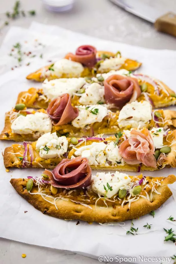 Angled shot of Butternut Squash Flatbread Pizza with Goat Cheese, Prosciutto & Pumpkin Seeds cut into long horizontal slices on a crinkled piece of parchment paper with a wood handled knife and small ramekin of salt blurred in the background