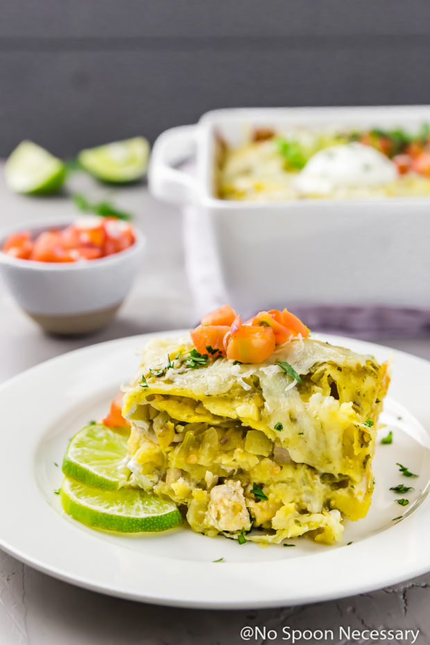 Straight on shot of a square slice of Easy Stacked Enchilada Suiza Bake on a white plate with slices of lime; there is a baking dish of Stacked Enchiladas, a small bowl of pico and lime wedges blurred in the background.
