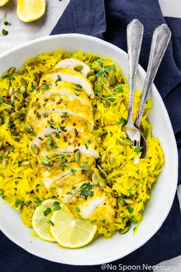 Overhead shot of Baked Curried Chicken & Spaghetti Squash in a neutral colored serving bowl and a spoon and fork inserted into the squash with a blue linen underneath the bowl and lemon wedges in the upper left corner of the shot.