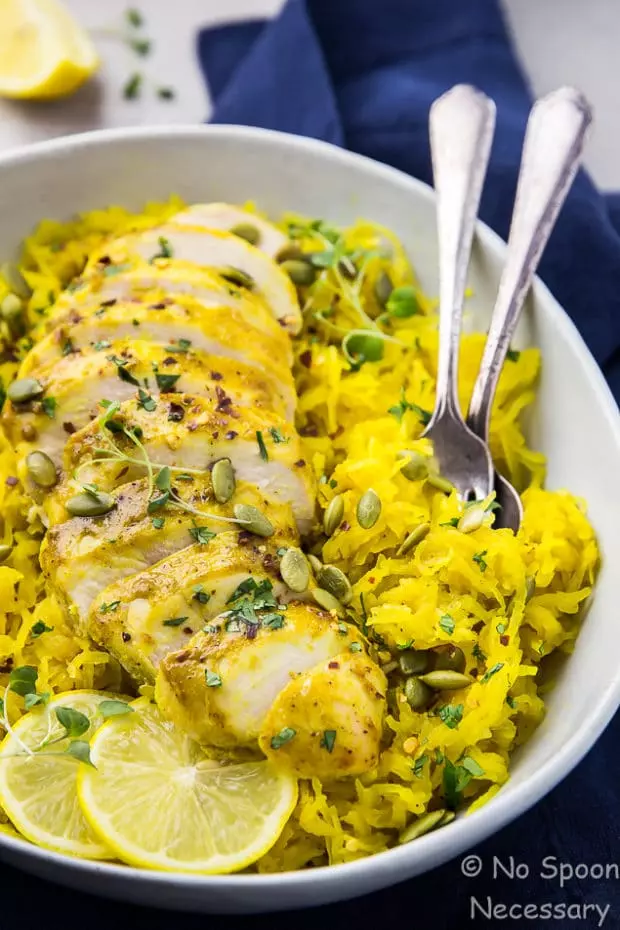 Angled shot of Baked Curried Chicken & Spaghetti Squash in a neutral colored serving bowl and a spoon and fork inserted into the squash with a blue linen underneath the bowl and lemon wedges in the upper left corner of the shot.