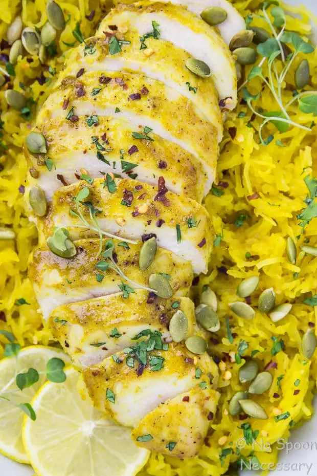 Overhead, up close shot of Baked Curried Chicken & Spaghetti Squash garnished with slices of lemon, pumpkin seeds and micro greens in a neutral colored serving bowl. 