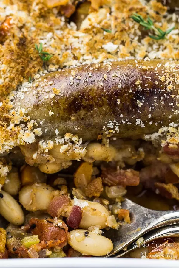 Up finish, angled shot of a Rooster Apple Sausage Hasty Cassoulet in a white baking dish with serving spoons tucked into the dish under the beans.