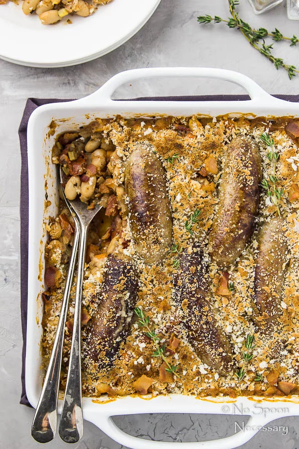 Overhead shot of a Rooster Apple Sausage Hasty Cassoulet in a white baking dish with serving spoons tucked into the dish and a plate of cassoulet, new thyme and salt and pepper shakers tucked into the tip of the shot.