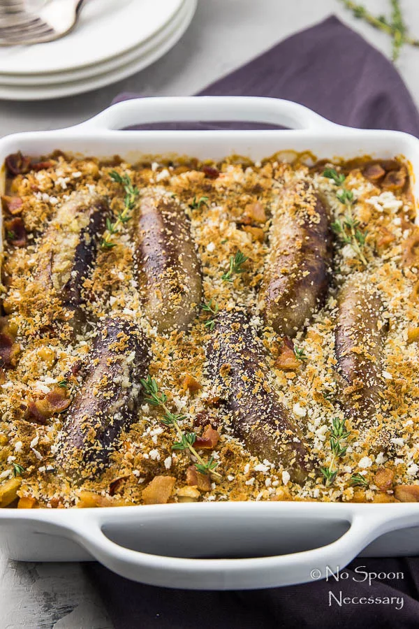 Angled shot of a Rooster Apple Sausage Hasty Cassoulet in a white baking dish with a deep crimson linen under the dish and a stack of plates with forks blurred on the benefit of the dish.