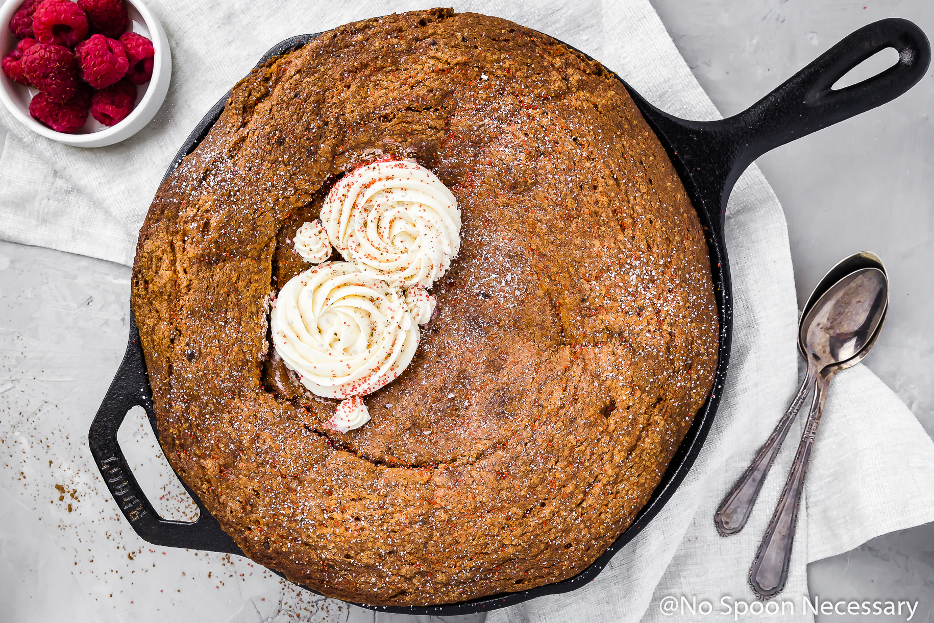 Overhead shot of a Chocolate Velvet Skillet Cake decorated with two "roses" of Cream Cheese Frosting on a gray surface and neural linen, with spoons and a ramekin of raspberries in the corner of the shot.