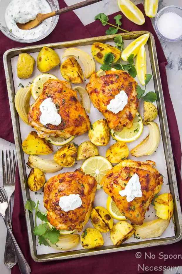 Overhead shot of a sheet pan of Harissa Chicken with Onions & Potatoes topped with herbed yogurt on a red linen with a fork and spoon in the bottom corner and lemon wedges, ramekin of salt, and a glass bowl of yogurt in the upper corners of the shot.