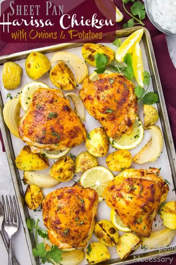 Overhead shot of a sheet pan of Harissa Chicken with Onions & Potatoes on a red linen with a fork and spoon in the bottom corner and a glass bowl of yogurt in the upper corner of the shot.