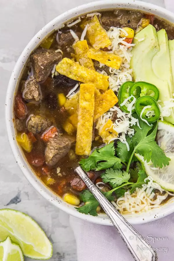 Overhead shot of Slow Cooker Beef Fajita Soup garnished with sliced avocado, cilantro, jalapenos, lime slices and tortilla strips with a spoon inserted into the soup, and a purple linen under the bowl and lime wedges in the corner of the shot.
