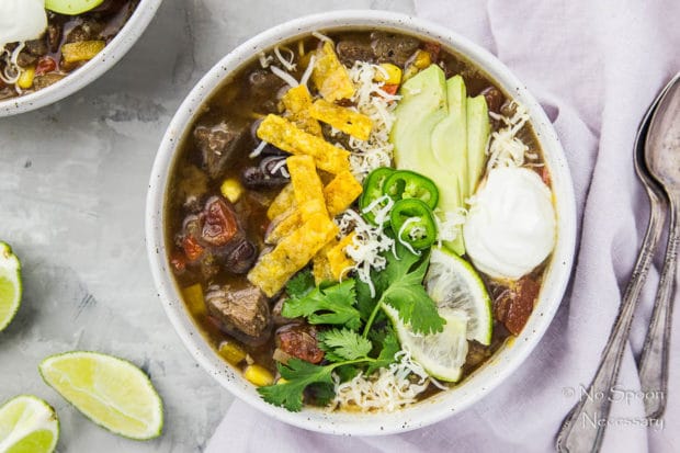 Overhead shot of Slow Cooker Beef Fajita Soup garnished with sliced avocado, cilantro, jalapenos, lime slices, tortilla strips and a dollop of sour cream; with a purple linen, spoons, lime wedges and another bowl of soup arranged around the bowl.