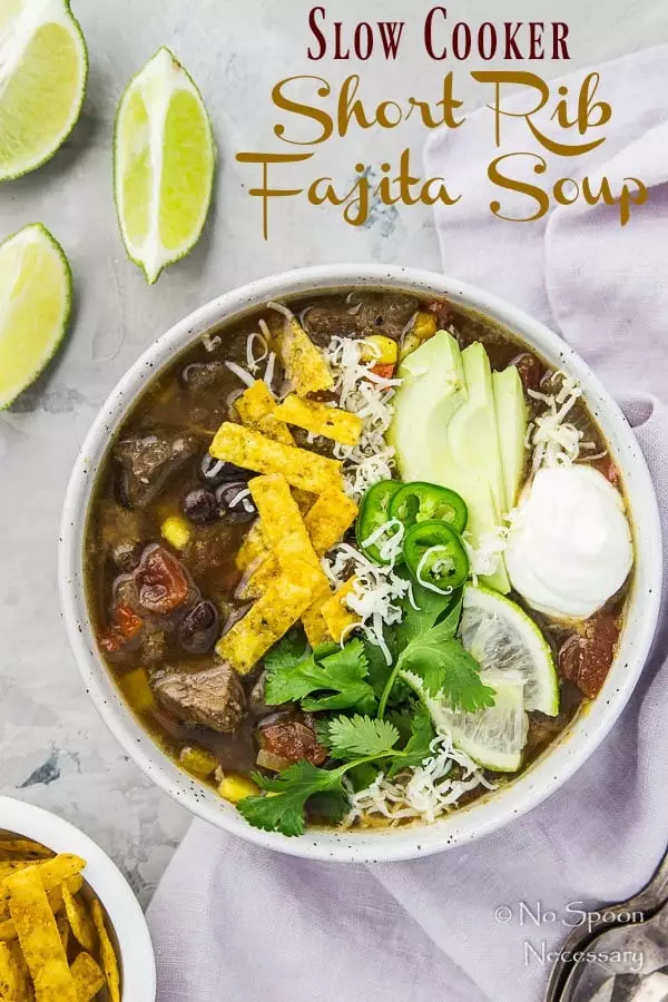 Overhead shot of Slow Cooker Beef Fajita Soup garnished with sliced avocado, cilantro, jalapenos, lime slices, tortilla strips and a dollop of sour cream; with a purple linen, spoons, lime wedges and ramekin of tortilla strips arranged around the bowl.