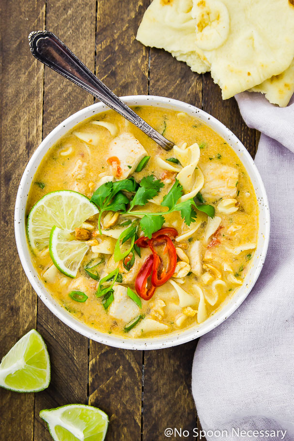 Overhead shot of a white bowl containing Slow Cooker Thai Style Chicken Noodle Soup garnished with cilantro, lime wedges, sliced red chilies and a spoon tucked into the soup; on a wood table with torn naan bread, lime wedges and a pale purple linen surrounding the bowl