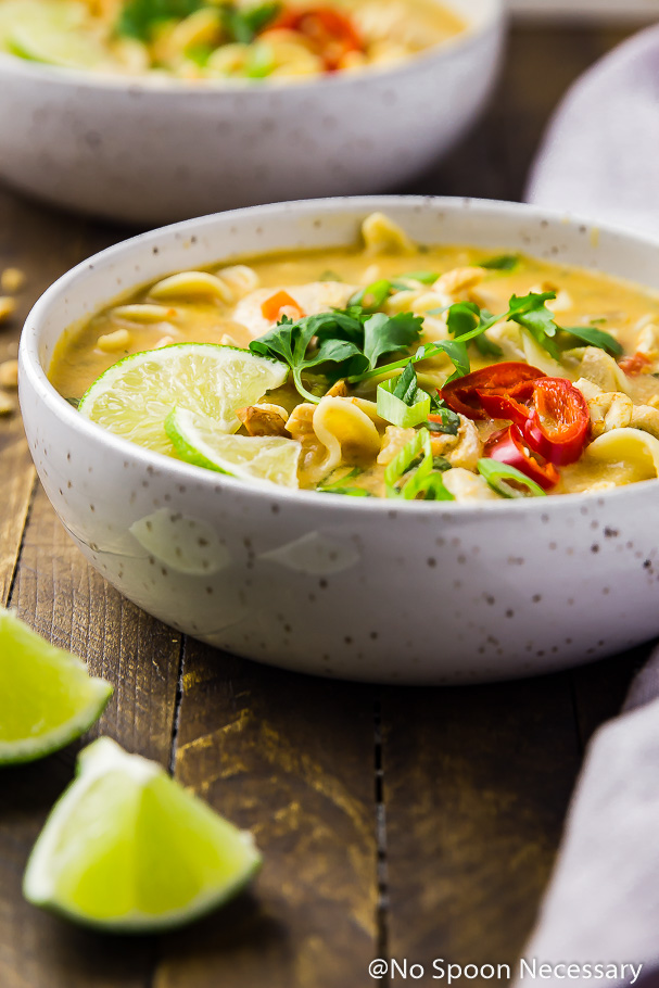 Straight on shot of a white bowl containing Slow Cooker Thai Style Chicken Noodle Soup garnished with cilantro, lime wedges and sliced red chilies on a wood table with lime wedges in front of the bowl and a second bowl of soup and a pale purple linen blurred in the background