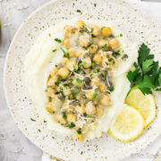 Overhead photo of Chickpea Piccata served over Cauliflower Mash on a speckled beige plate with lemon slices and parsley; with wedges of lemon and a small block of parmesan off to the sides of the plate.