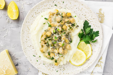 Overhead photo of Chickpea Piccata served over Cauliflower Mash on a speckled beige plate with lemon slices and parsley; with wedges of lemon and a small block of parmesan off to the sides of the plate.