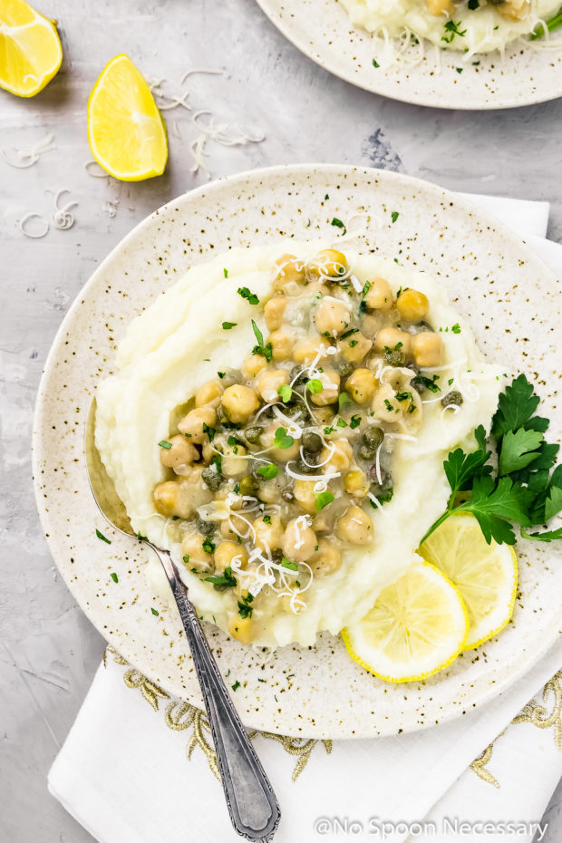 Overhead shot of Chickpea Piccata with Cauliflower Mash on a speckled beige plate with lemon slices and parsley; with another plate of piccata and lemon wedges in the corner of the shot.