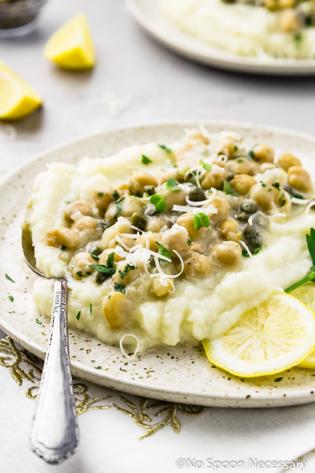 45 degree angle shot of Chickpea Piccata with Cauliflower Mash on a speckled beige plate with a spoon inserted into the 'potatoes' and another plate of piccata, lemon wedges and a small ramekin of capers blurred in the background.