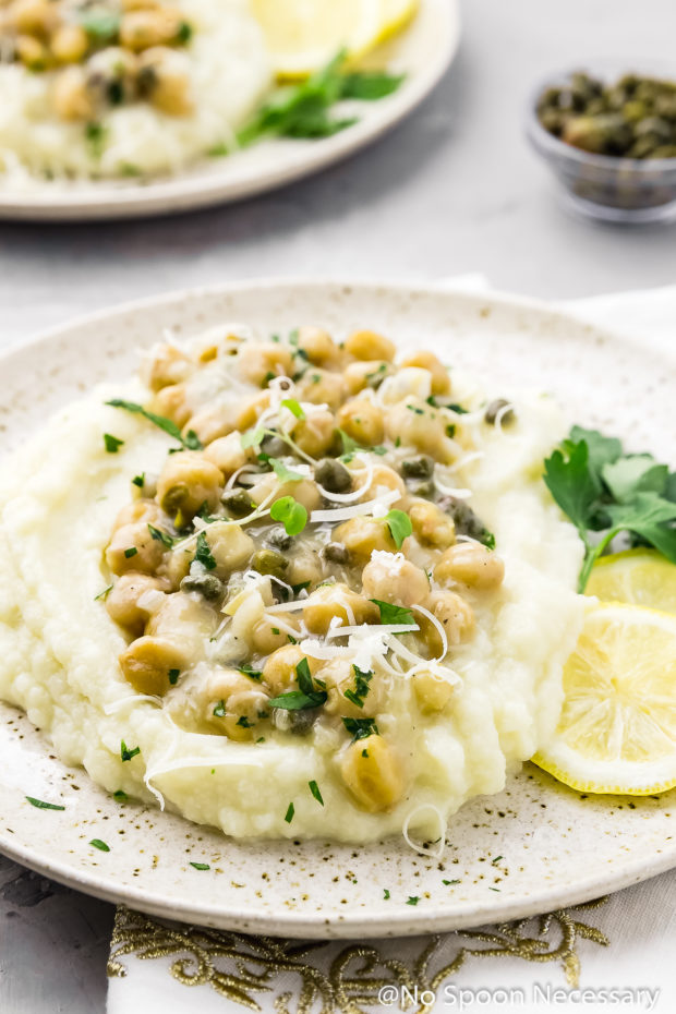 45 degree angle shot of Chickpea Piccata with Cauliflower Mock Mashed 'Potatoes' on a speckled beige plate with lemon slices and parsley; with another plate of piccata and a small ramekin of capers blurred in the background.