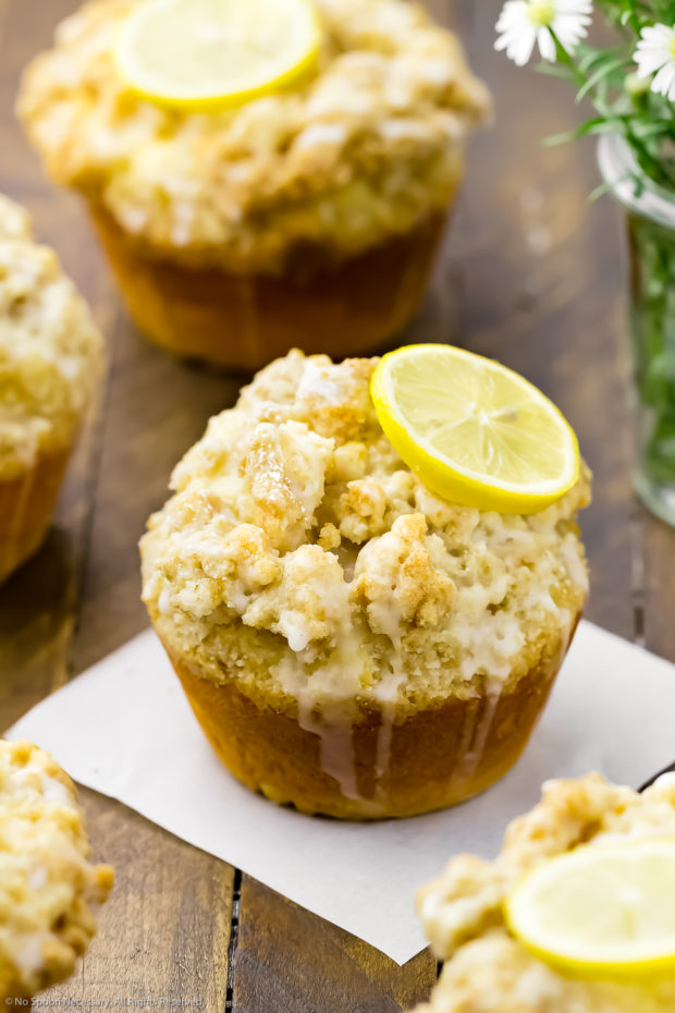 Angled shot of Glazed Lemon Muffins on a wooden board topped with lemon slices with the focus of the shot on one individual muffin on top of a small piece of parchment paper.