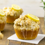 Straight on shot of Glazed Jumbo Lemon Crumb Muffins on a wooden board with spring flowers and lemon halves in the background and the focus of the shot on one individual muffin.