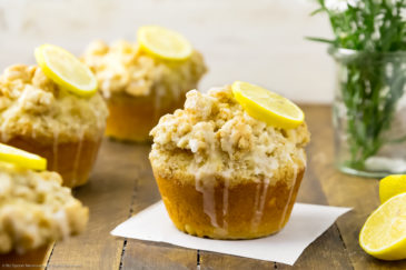 Straight on shot of Glazed Jumbo Lemon Crumb Muffins on a wooden board with spring flowers and lemon halves in the background and the focus of the shot on one individual muffin.