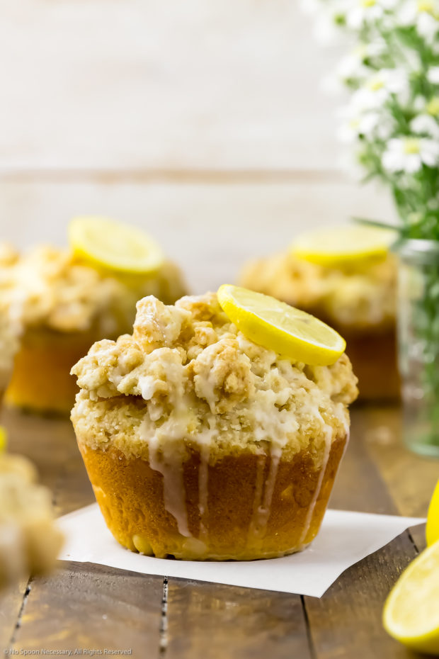 Straight on shot of Glazed Lemon Muffins on a wooden board with spring flowers in the background and the focus of the shot on one individual muffin.