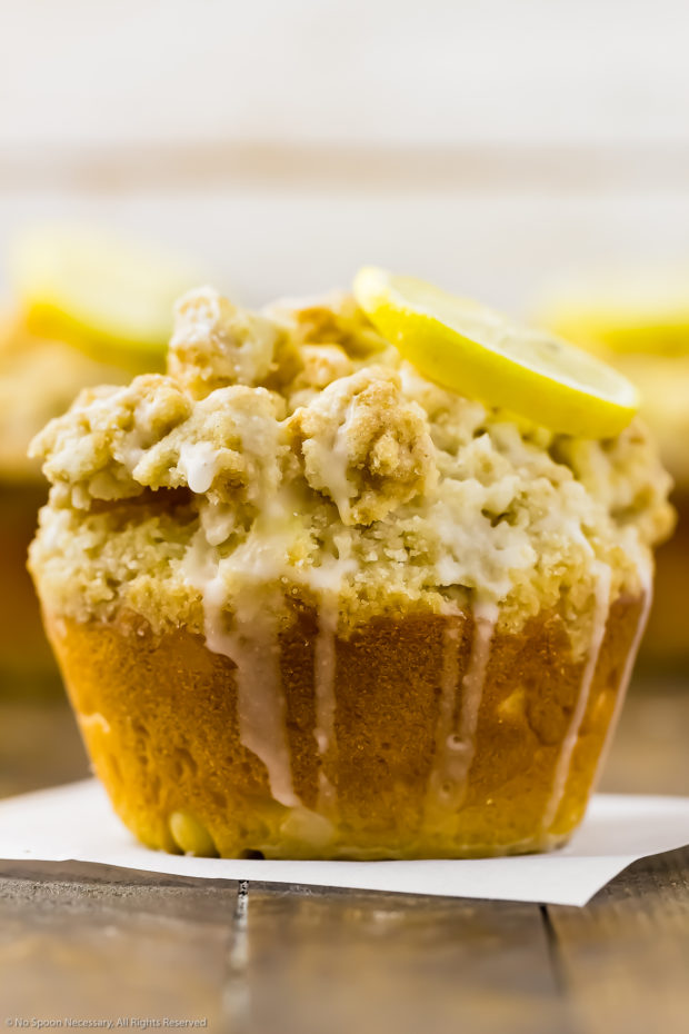 Straight on shot of a Glazed Lemon Crumb Muffins topped with a lemon slice on a wooden board with more muffins blurred in the background.